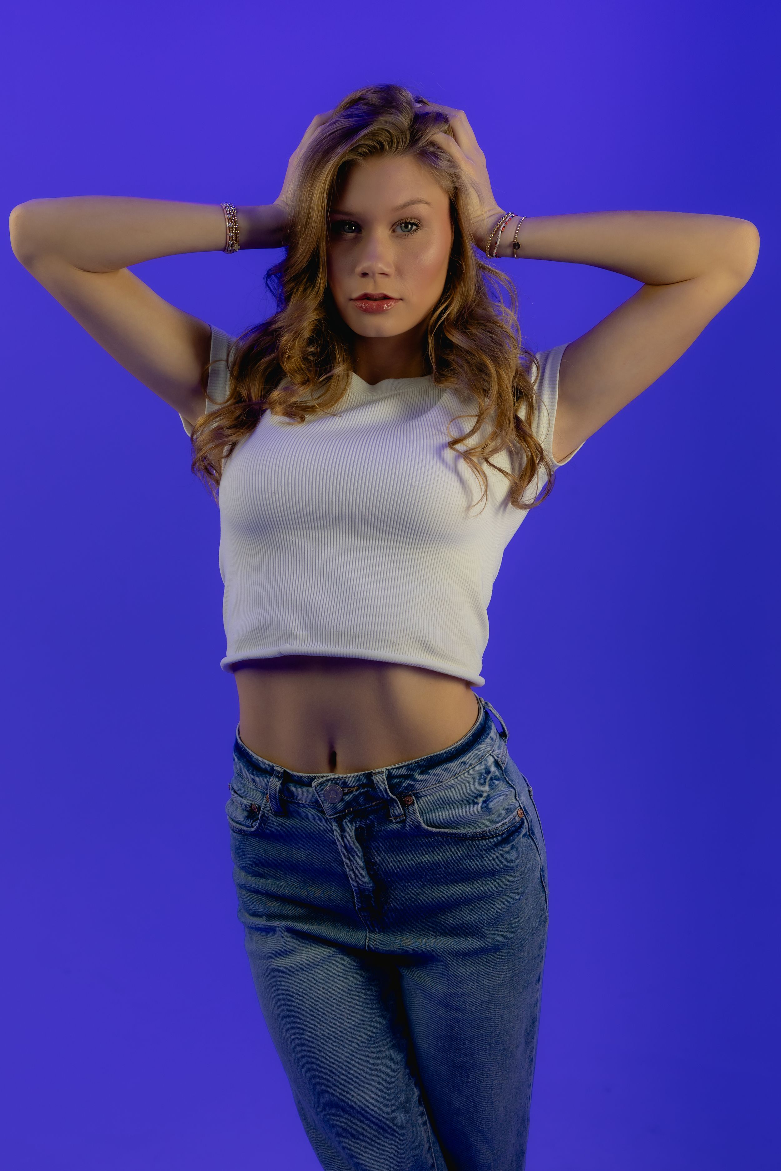 A model poses with hands behind head wearing a white crop top and blue jeans against a vibrant blue background.