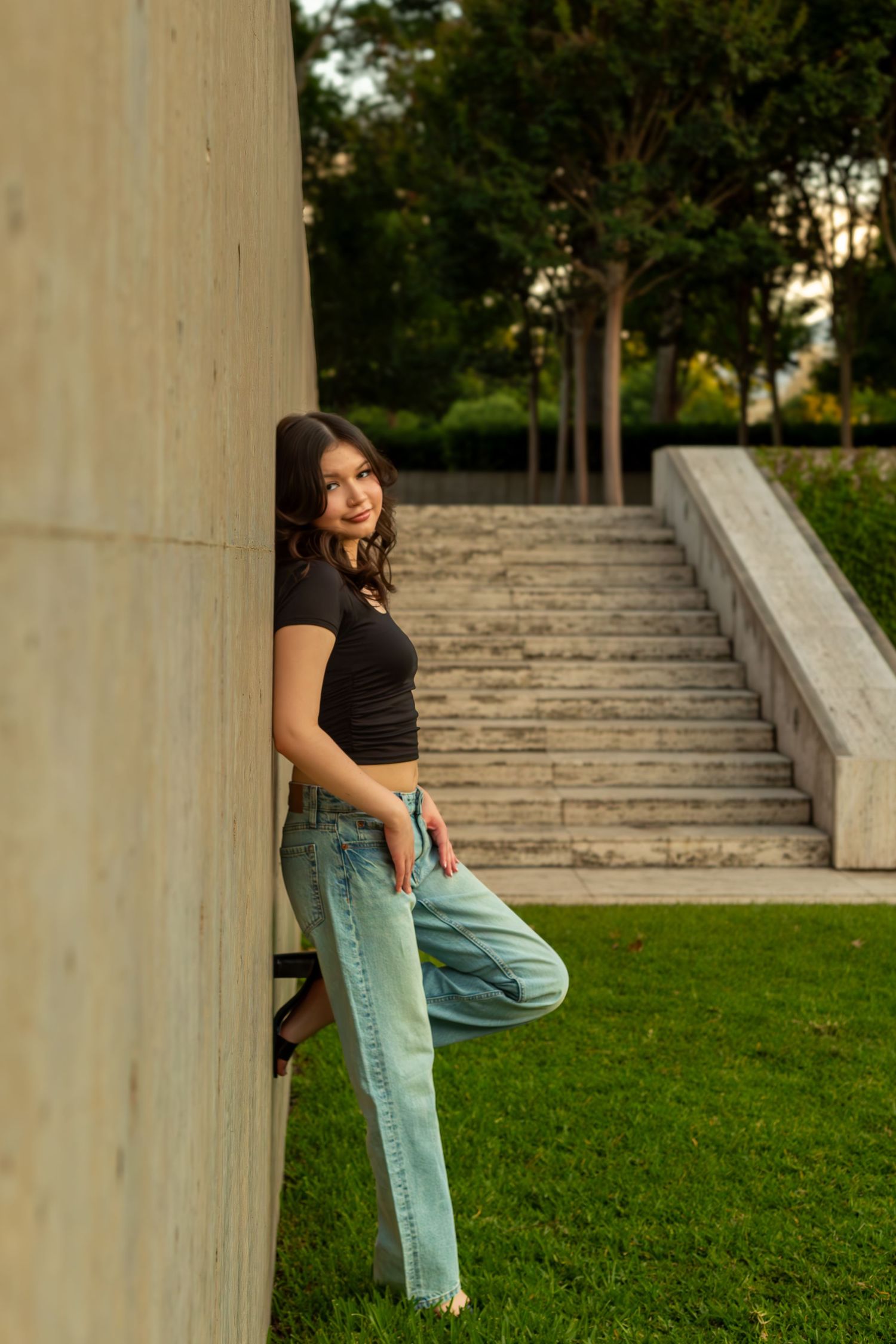 A person in casual attire leans against a concrete pillar near stone steps and grassy area during golden hour.