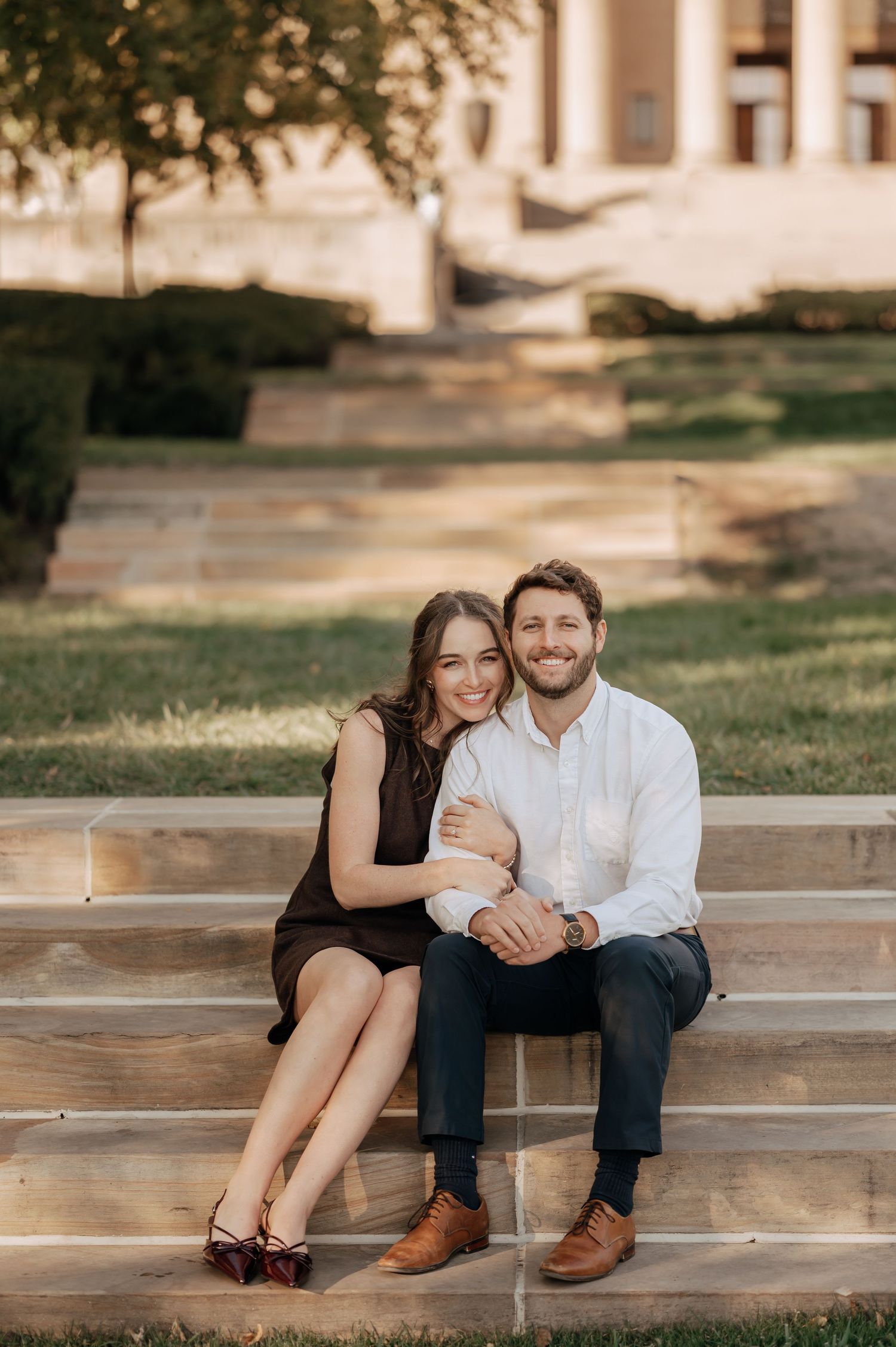 Smiling couple sitting on outdoor steps at golden hour, woman in black dress leaning on man.