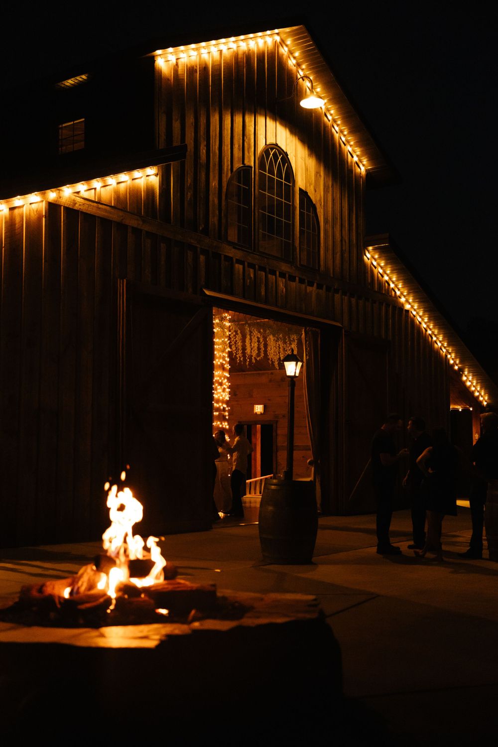 Warm string lights illuminate a rustic barn exterior while a bonfire glows in the foreground at night.