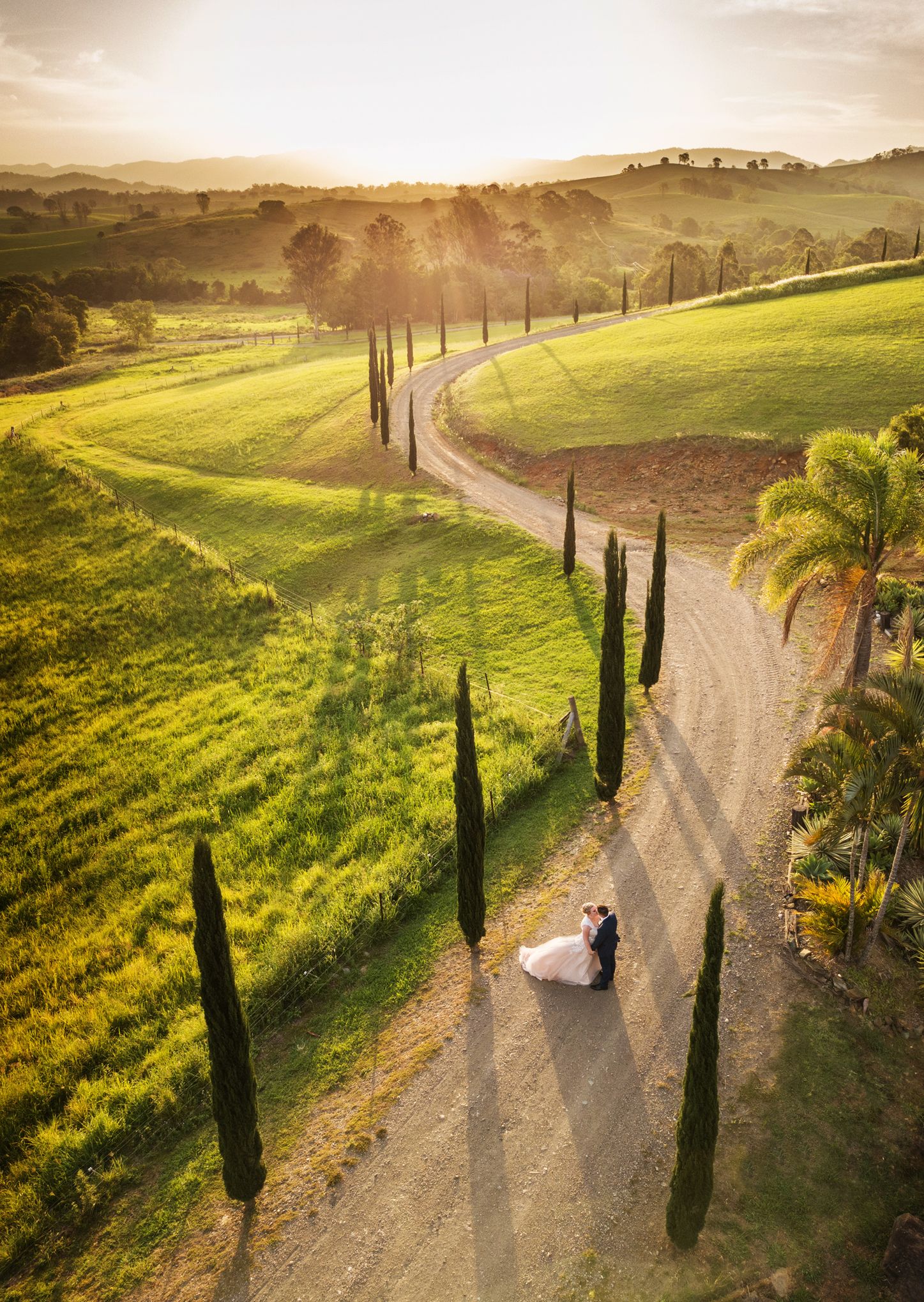 Aerial view of bride and groom embracing on a winding driveway surrounded by lush green hills at sunset.