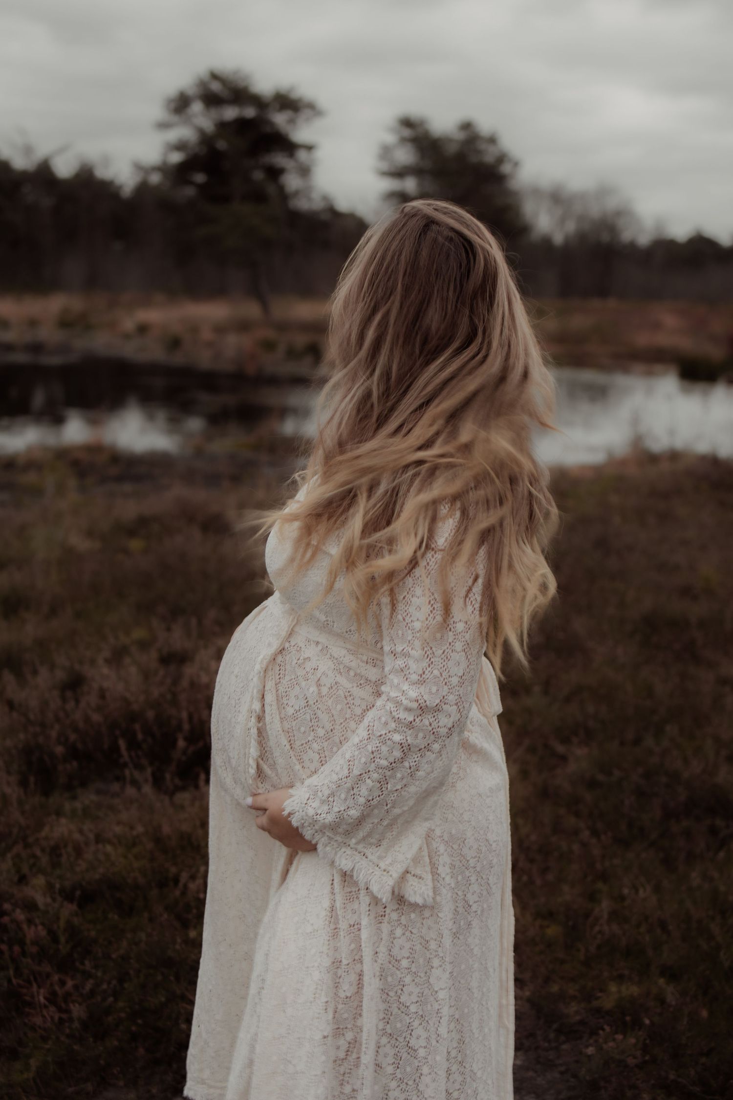 Ethereal maternity photo in flowing white dress by tranquil lake at sunset with soft sepia tones.