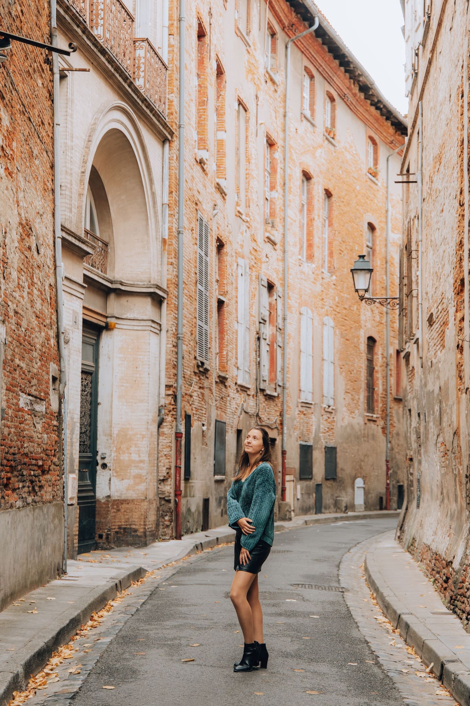 Woman in green sweater and black skirt standing on a narrow European brick building street.