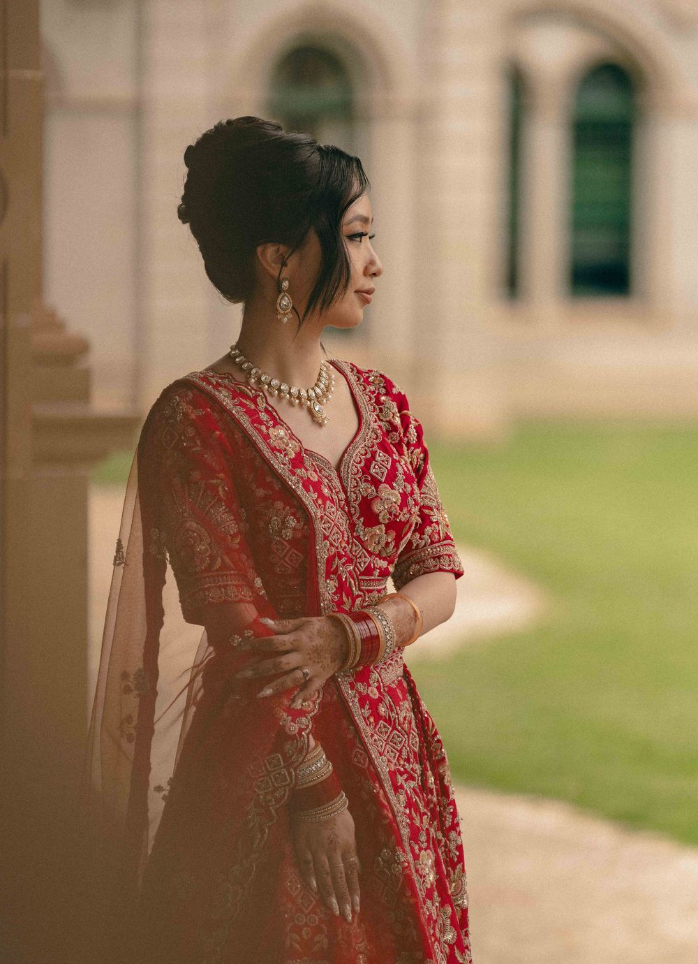 A person in an elegant red embroidered sari poses gracefully against a classic architectural backdrop.