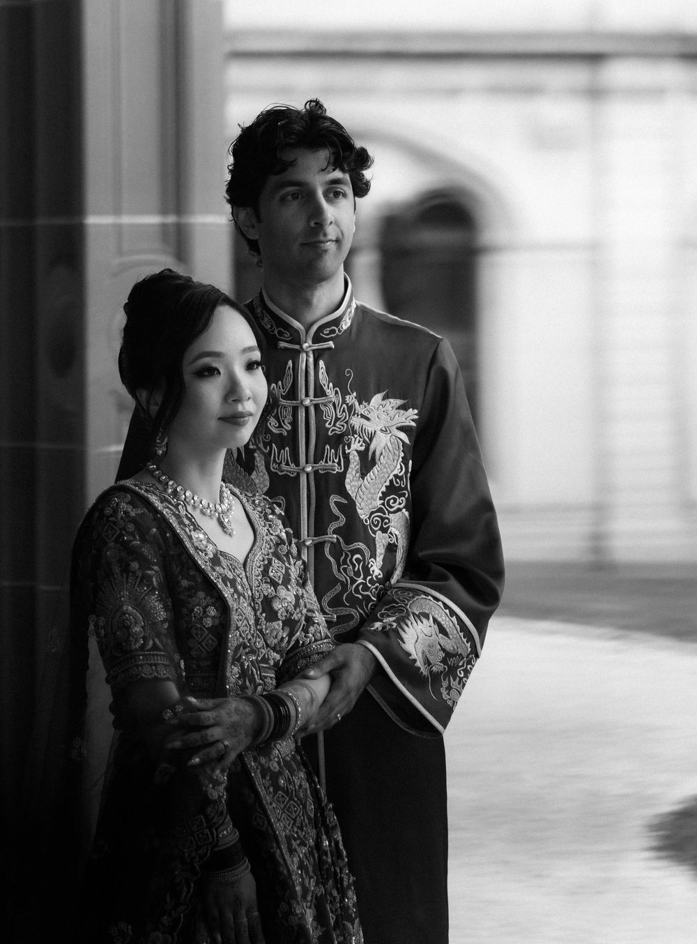 A black and white portrait of a couple in traditional Asian formal attire with embroidered details standing by a window.