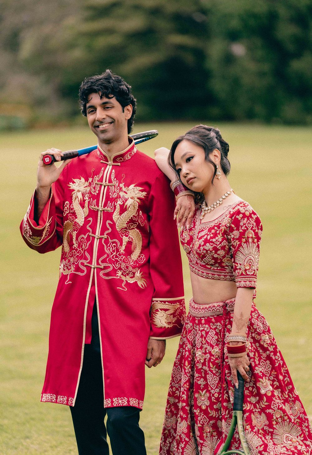 Couple in matching red and gold traditional attire, man in Chinese jacket, woman in Indian lehenga, posing outdoors.