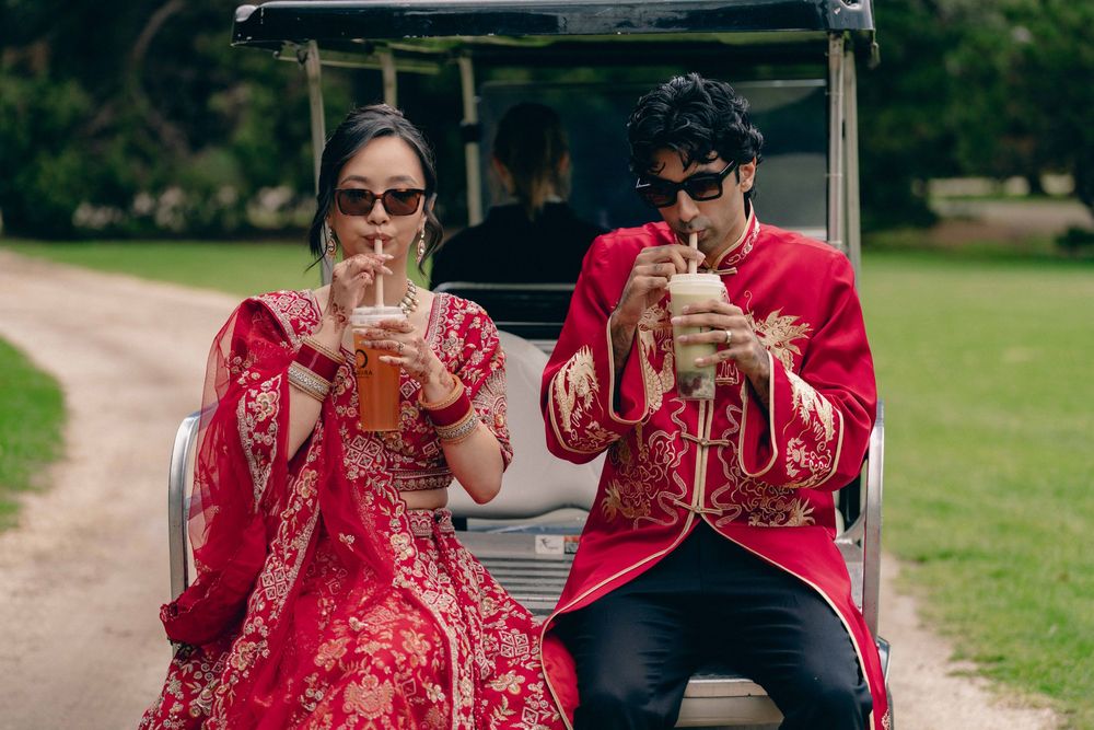 Couple in red traditional Indian attire sipping drinks on a golf cart along a scenic tree-lined path.