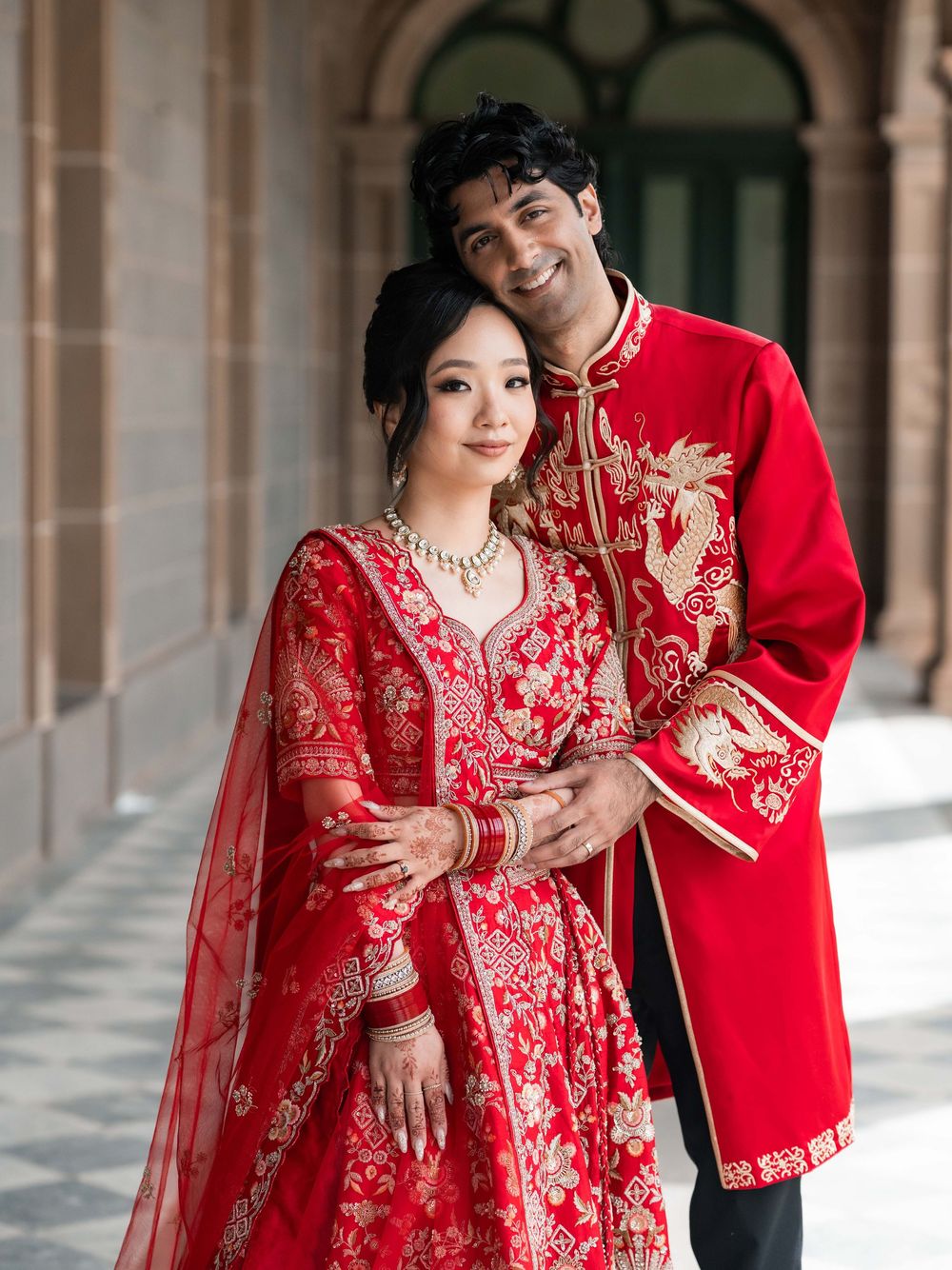 A couple in traditional red wedding attire with gold embroidery poses together in an elegant arched hallway.