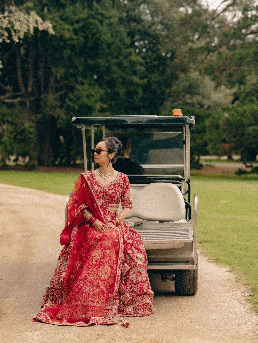 A person in a red embroidered bridal lehenga sits elegantly in a golf cart on a tree-lined path.
