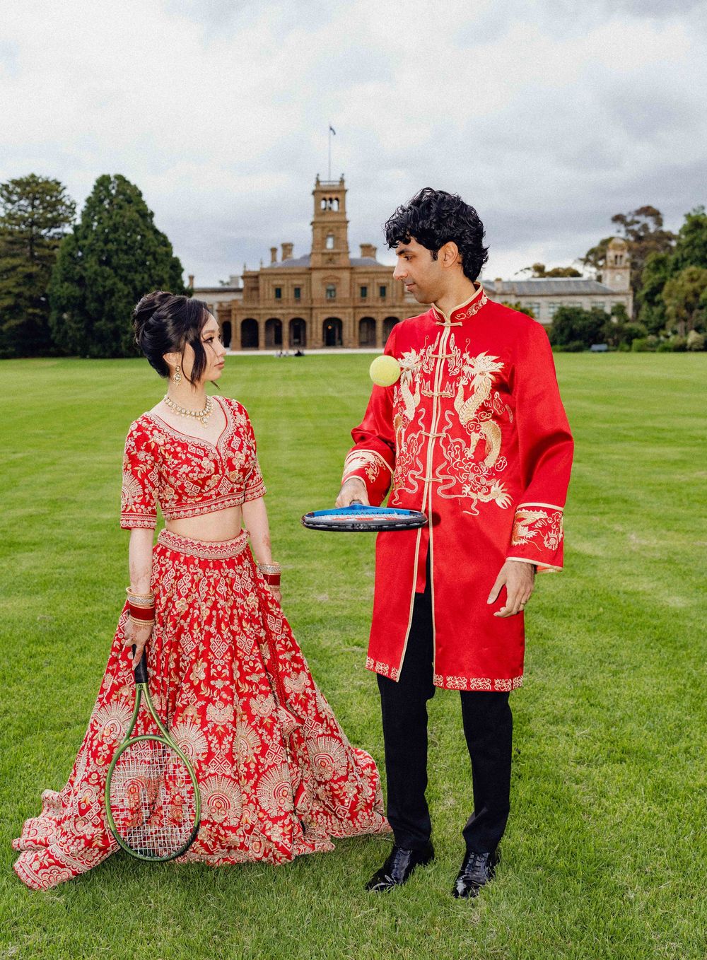 Couple in matching red traditional outfits holding badminton rackets on green lawn with historic building backdrop.