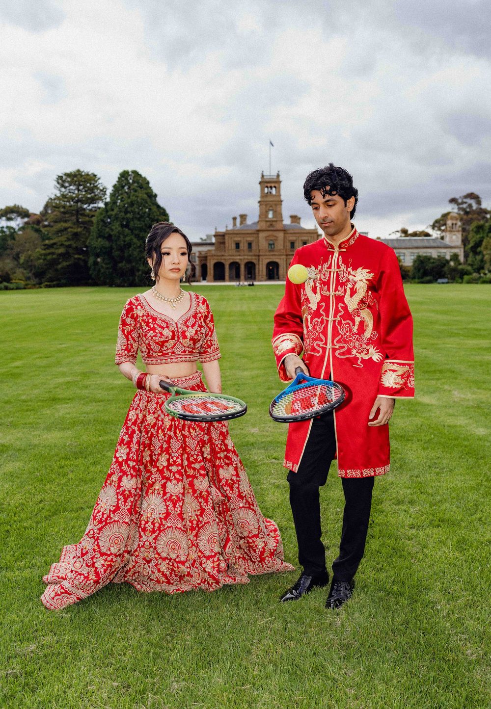 Couple in matching red and gold traditional South Asian attire holding badminton rackets on green lawn before grand estate.