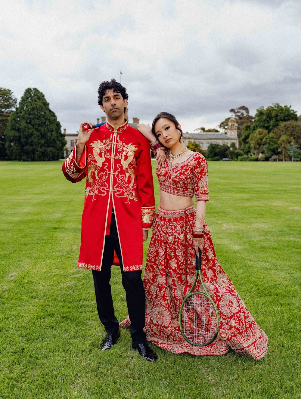 Couple in red traditional South Asian and Chinese wedding attire posing on green lawn holding tennis racket and roses.