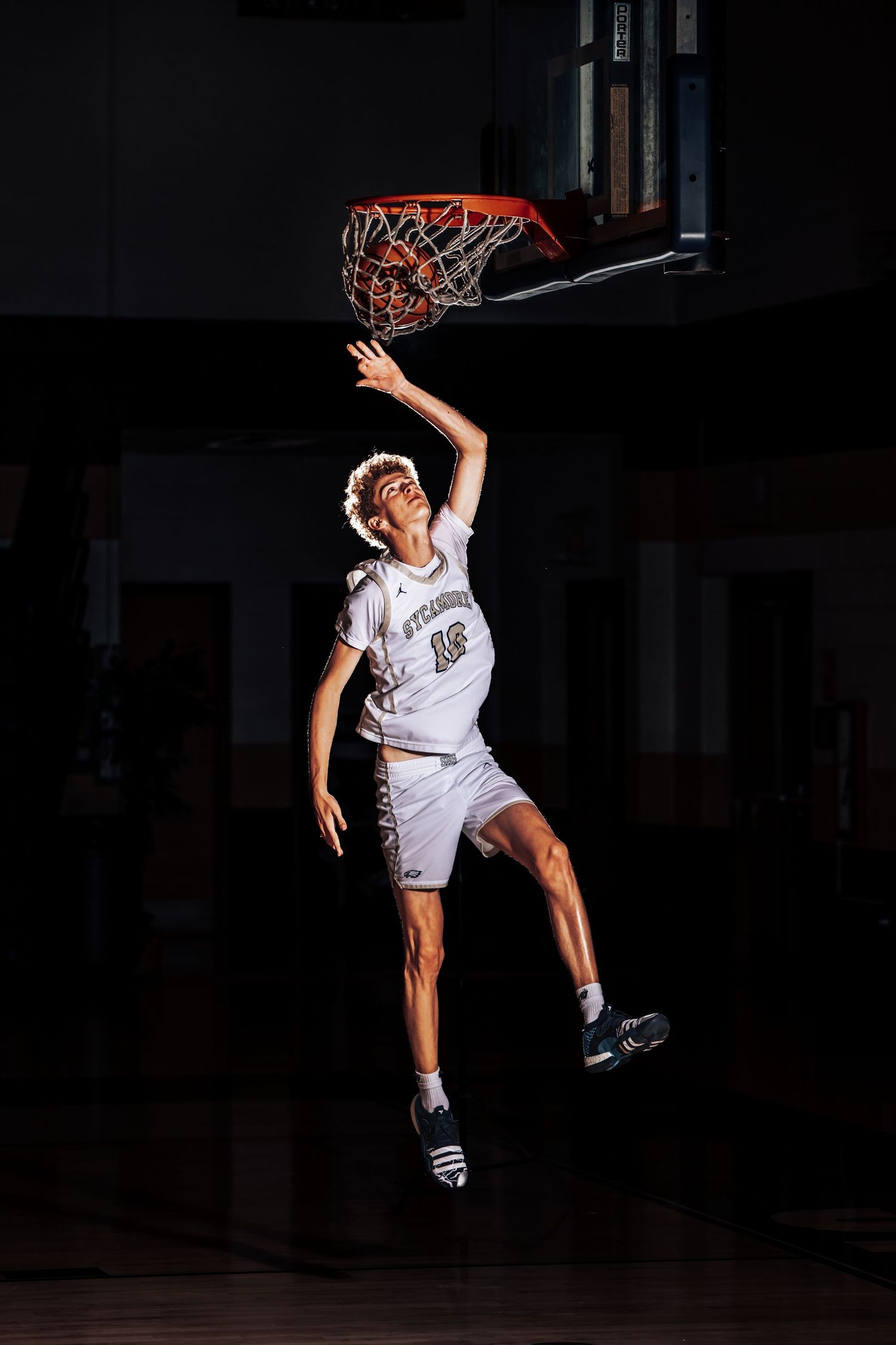 Basketball player in white jersey performs dramatic slam dunk sequence in dark indoor court setting.