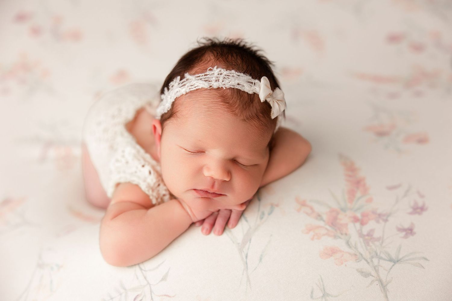 Sleeping newborn baby wearing white lace headband on pink floral backdrop.