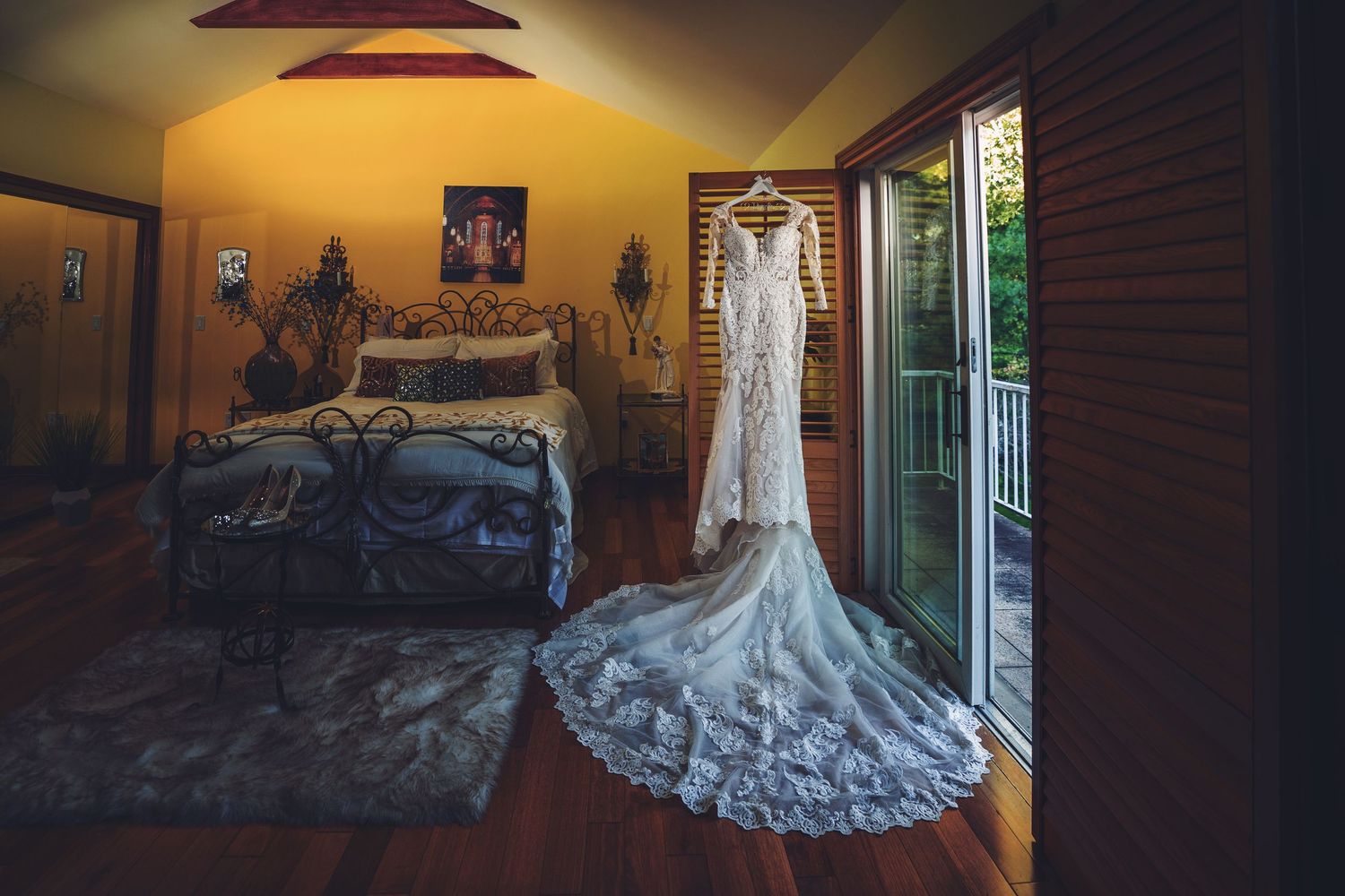 An elegant lace wedding dress hangs in a warmly lit bedroom with dramatic lighting.