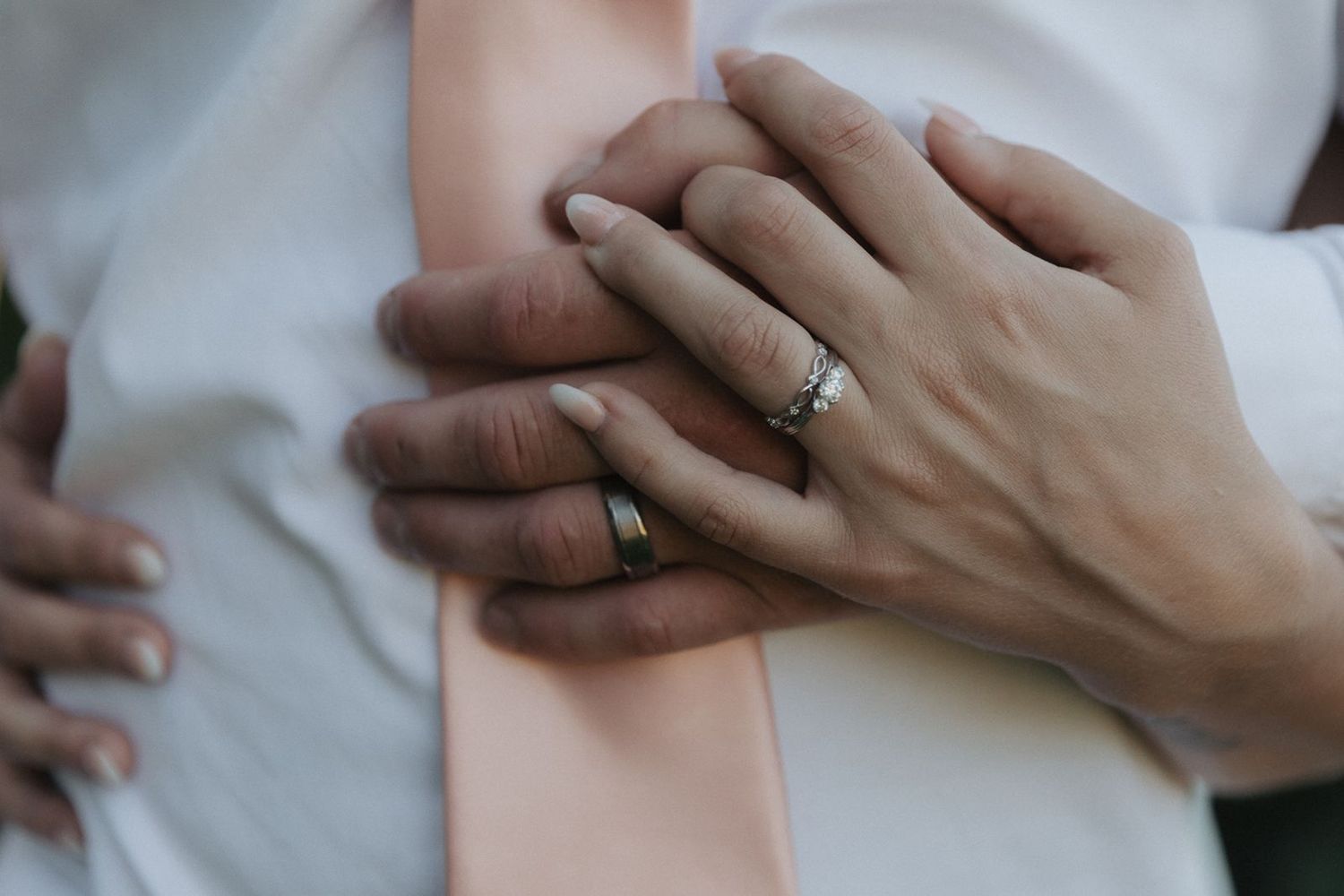 Close-up of newlyweds hands with wedding rings resting on grooms white shirt and blush pink tie.
