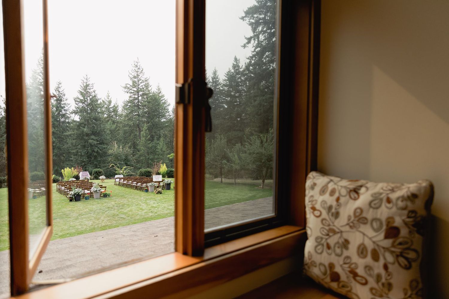 View through wooden-framed window overlooking misty outdoor wedding ceremony setup among evergreen trees.