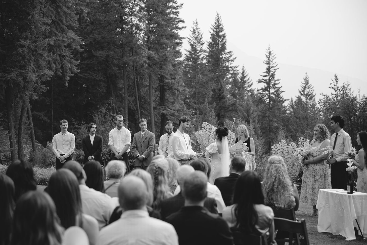 Black and white wide shot of outdoor wedding ceremony with guests seated among tall evergreen trees.