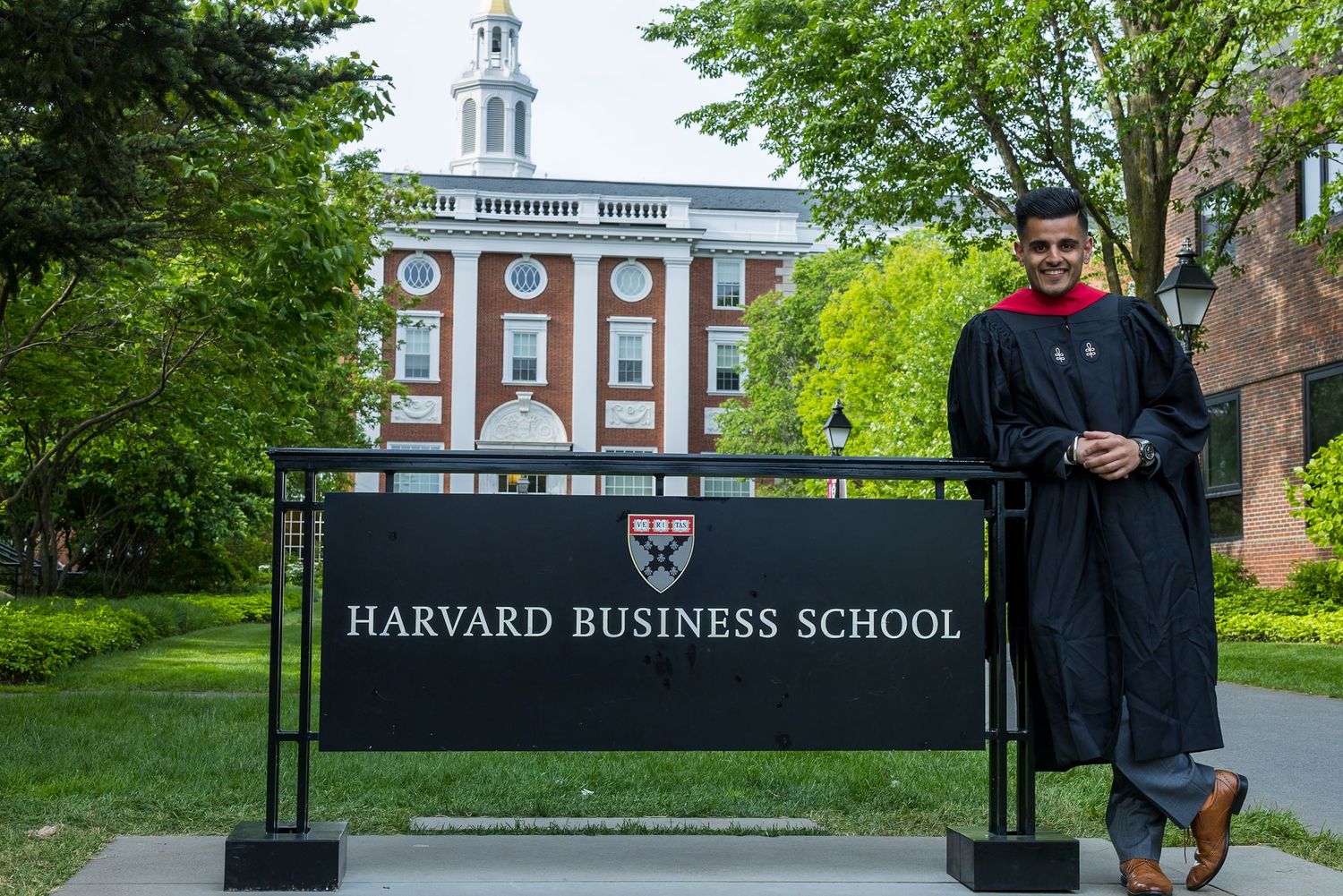 A graduate stands next to the Harvard Business School sign with a historic brick building and green trees in the background.