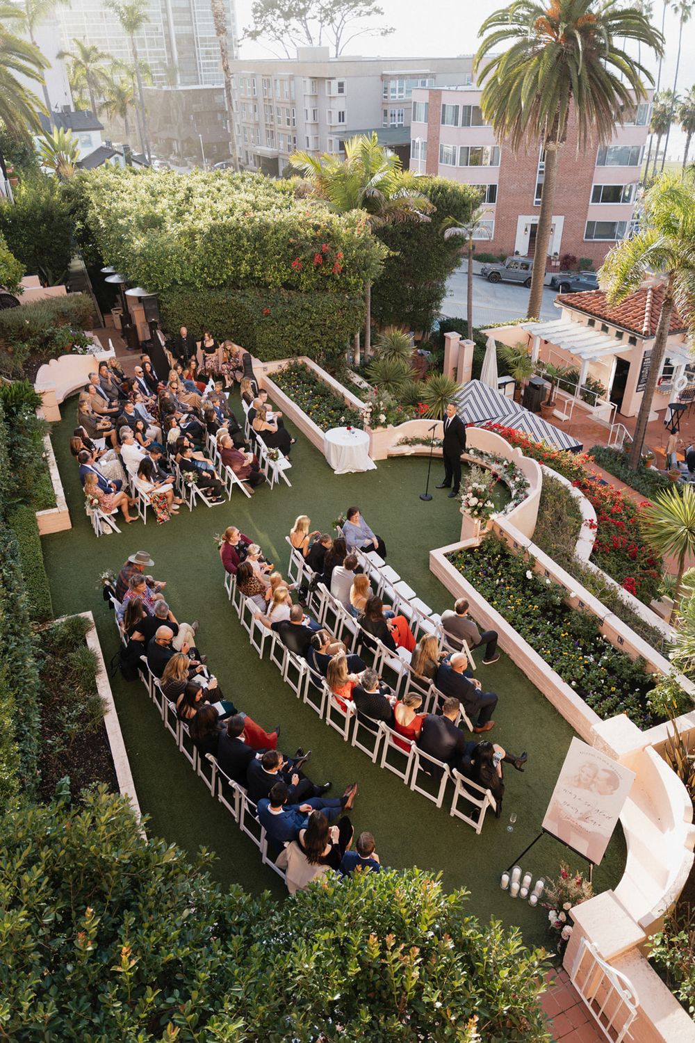 Aerial view of an outdoor garden wedding ceremony with white chairs arranged in rows and surrounded by lush greenery.