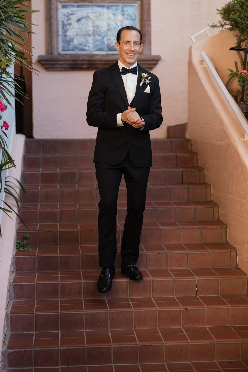 A person in a black tuxedo sits and poses on terracotta-colored outdoor steps near a beige stucco wall.