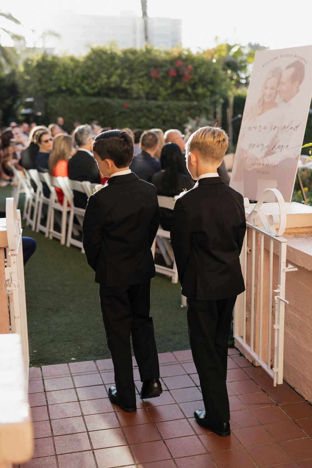 Young boys in formal black suits gather near white chairs at an outdoor wedding ceremony.