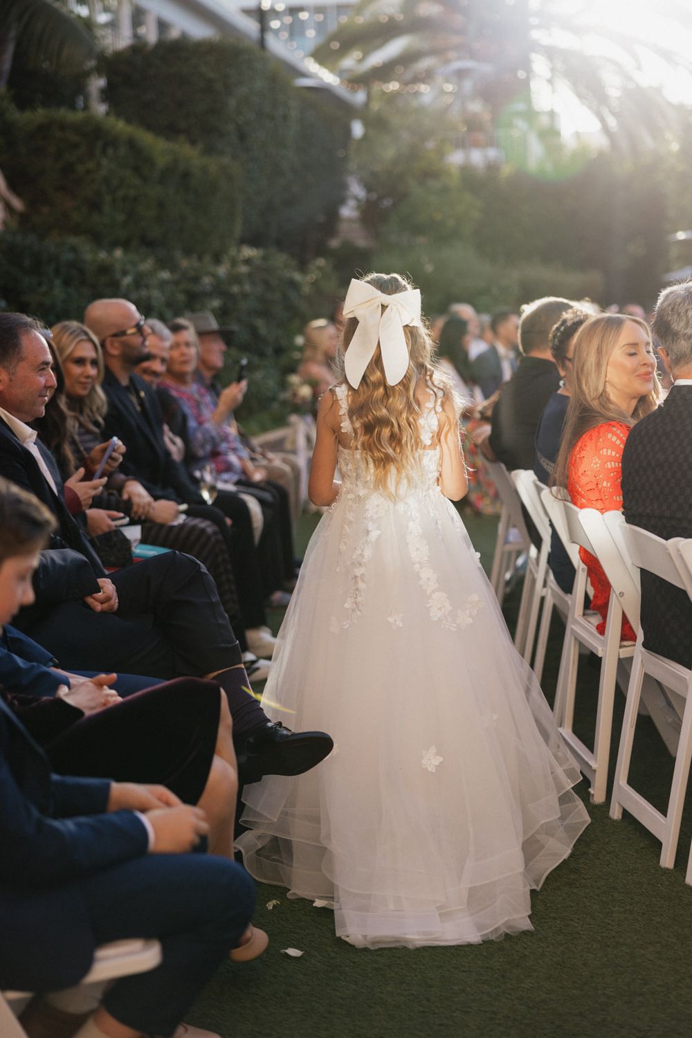A bride in a flowing white dress stands among wedding guests during an outdoor sunset ceremony.