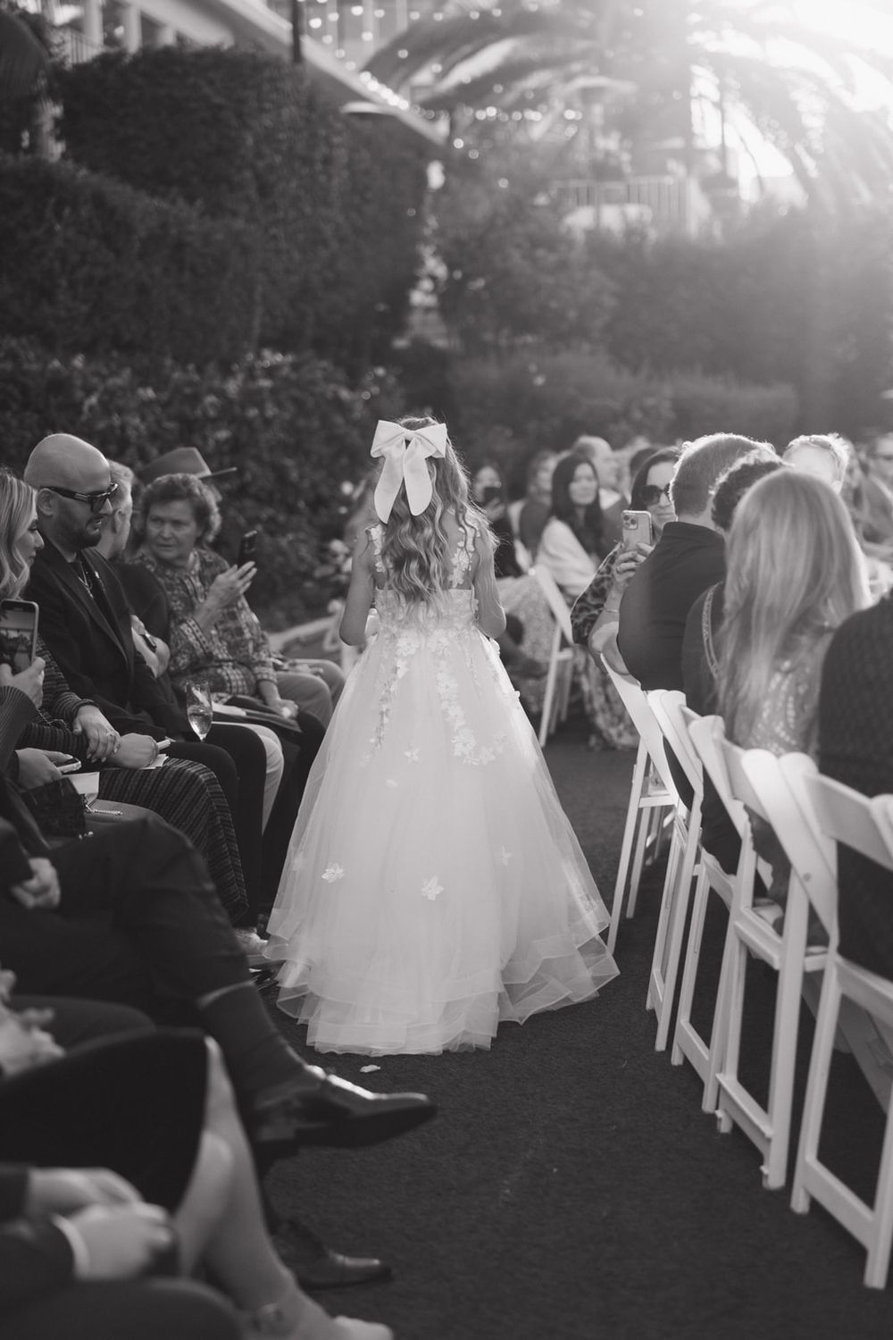 Wedding guests seated in white chairs watch as brides in flowing white gowns walk down an outdoor aisle at sunset.