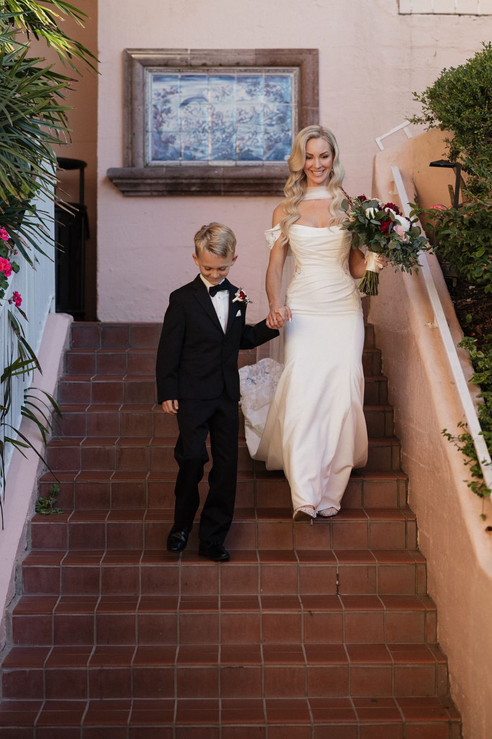 A young ring bearer in a black tuxedo escorts someone in a white wedding gown down terracotta steps at an outdoor venue.