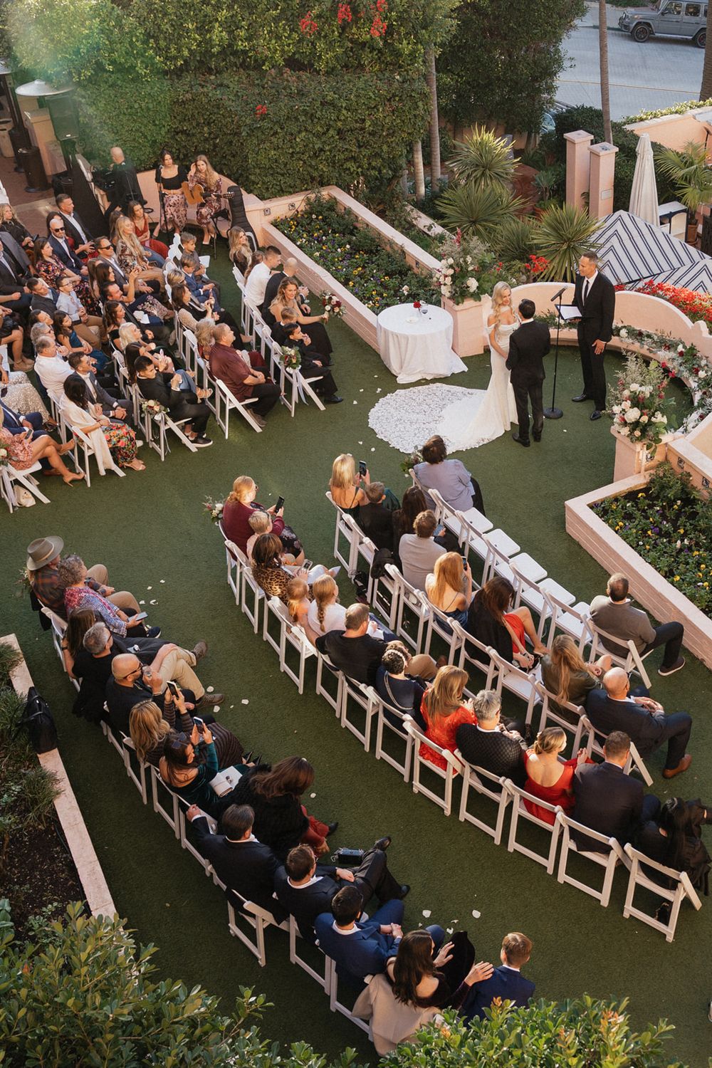 Wedding ceremony guests seated on white chairs in a garden setting during an outdoor celebration.