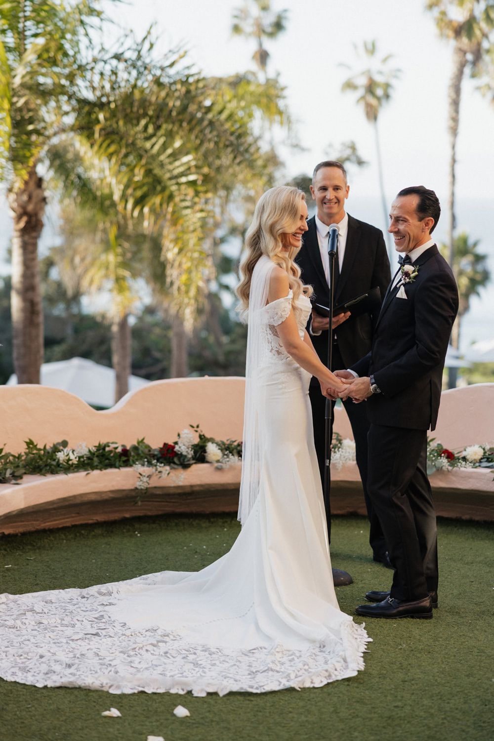 A wedding ceremony takes place on a terrace with palm trees in the background as a couple exchanges vows.