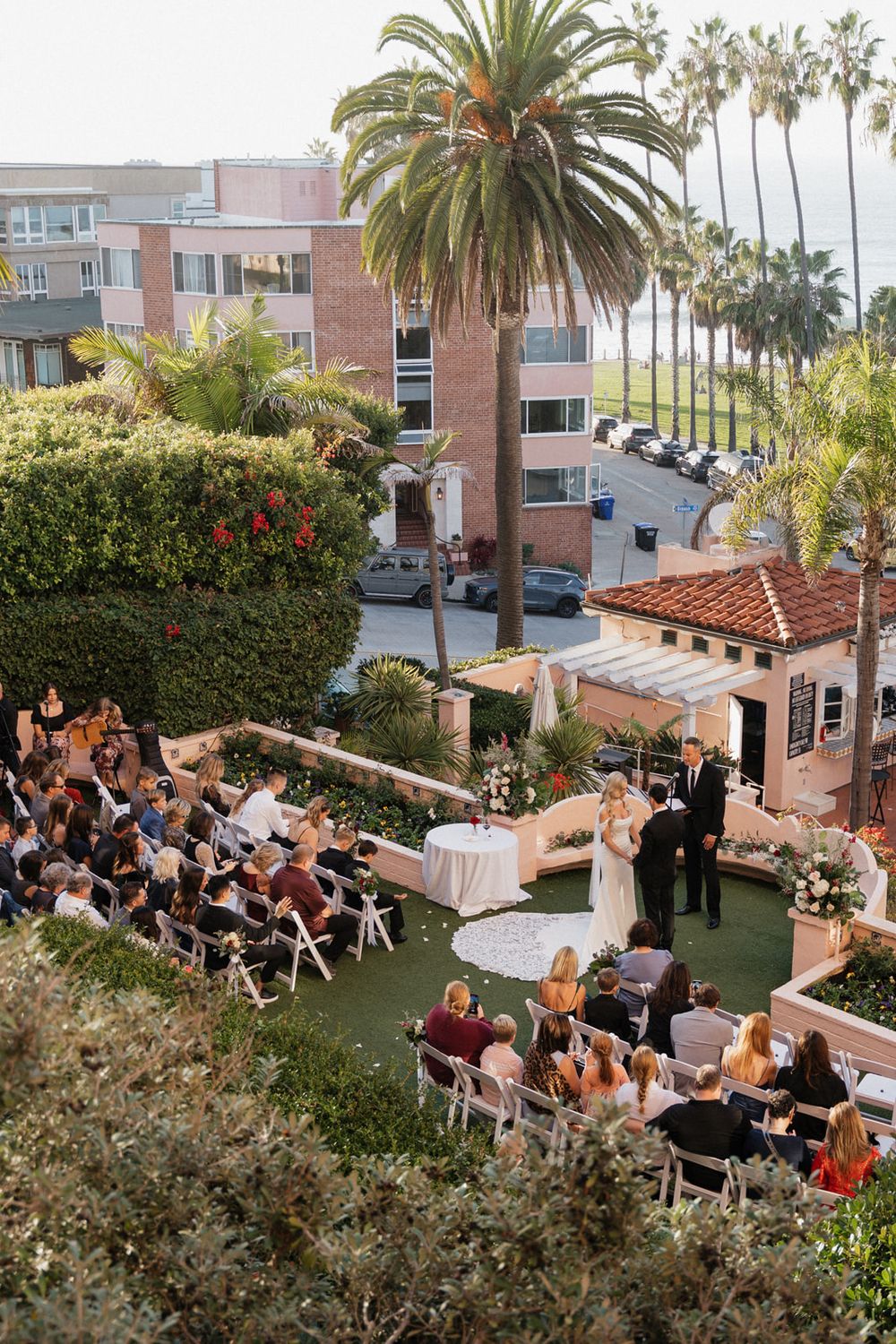 An aerial view of an outdoor wedding ceremony with guests seated on a lawn surrounded by palm trees and buildings.