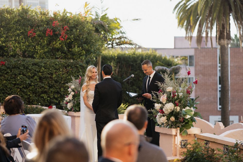 Panoramic view of an outdoor wedding ceremony with guests seated in rows of white chairs surrounded by palm trees.