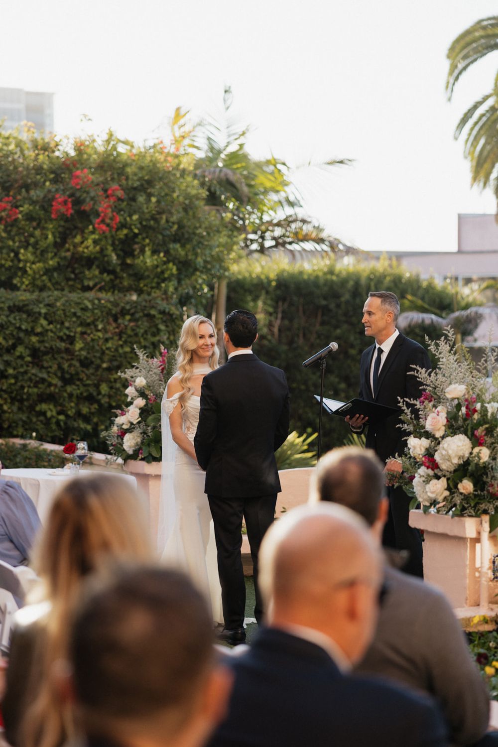 Outdoor wedding ceremony with guests watching the bride and groom exchange vows under floral arrangements and palm trees.