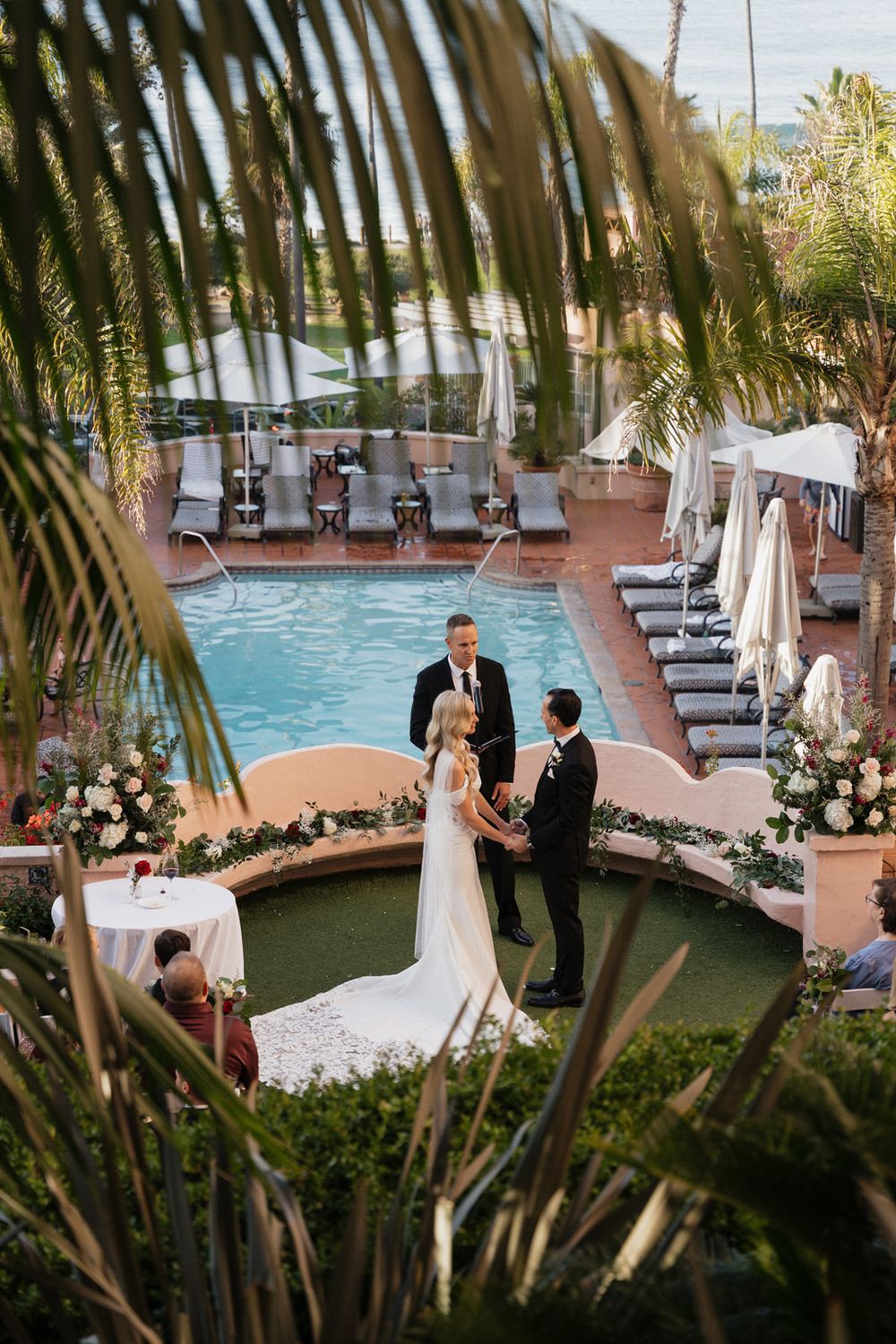 Wedding ceremony taking place by a pool with palm trees and lounge chairs in the tropical setting.