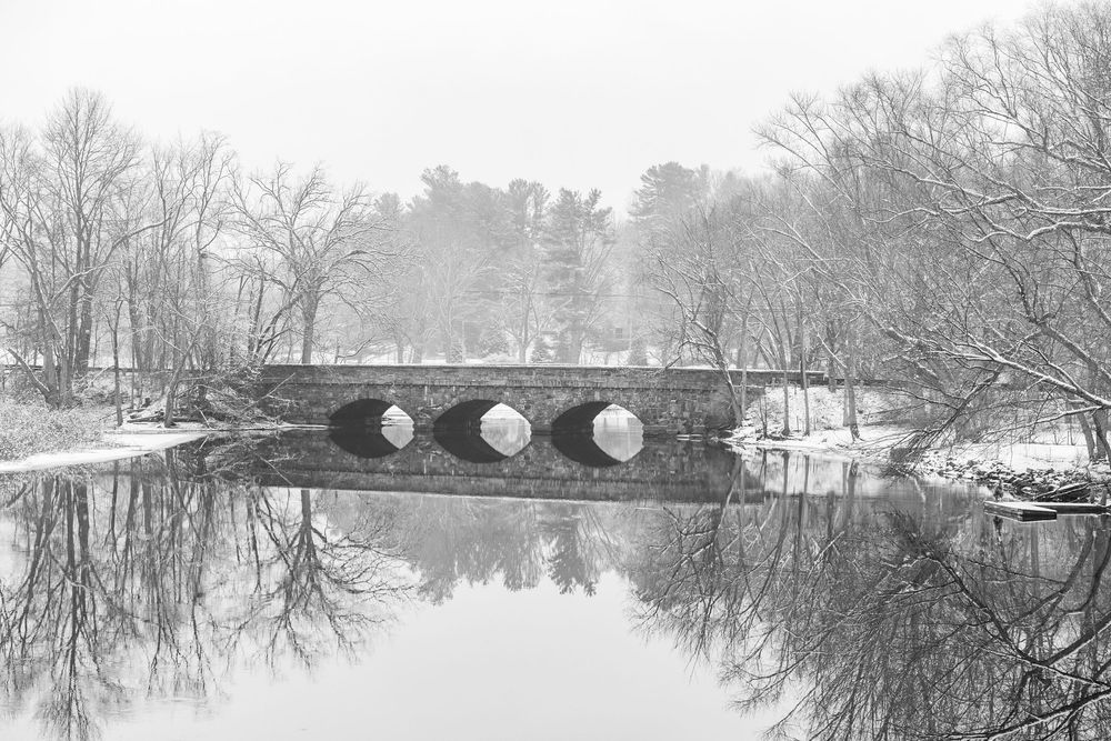 A panoramic winter landscape shows snow-covered trees and a stone bridge reflected on Concord River on a snowy day.