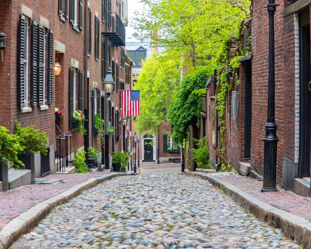 Historic cobblestone street with brick townhouses and American flag in Boston's Beacon Hill neighborhood on Acorn Street.