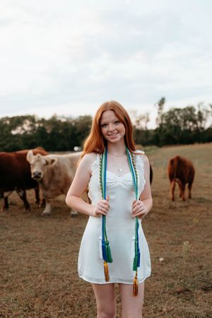 A person in a white top stands in a field with horses grazing in the background during golden hour.