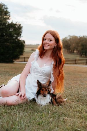 Person with long red hair wearing white dress lies in grass with small brown dog in rustic outdoor setting.