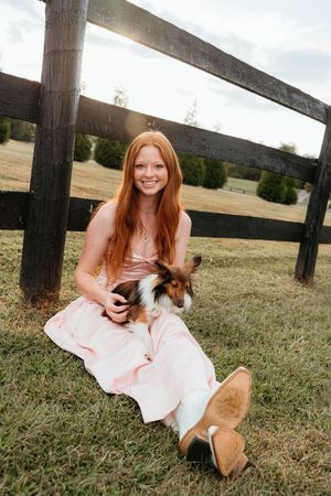 A person in a pink dress sits on grass near a wooden fence with a small brown and white dog.