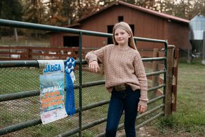 Person in brown sweater and jeans leaning against rustic wooden fence in outdoor setting.