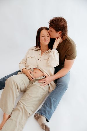 A man tenderly kisses a pregnant woman's cheek while cradling her baby bump in a bright studio setting.
