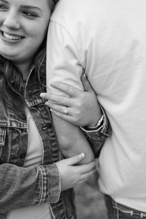 Black and white photo of smiling woman in denim jacket hugging man's arm, showing engagement ring on her finger.