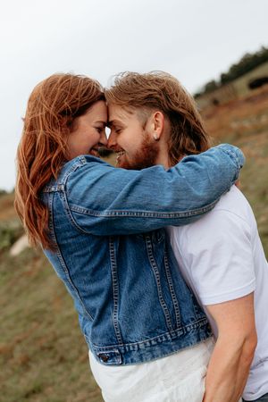 A romantic couple with long hair embraces outdoors, touching foreheads and smiling, woman in denim jacket, man in white tee.