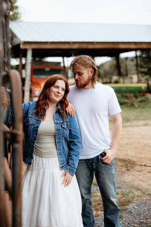 A young couple poses outdoors near a farm, woman in denim jacket and white dress, man in white tee and jeans.