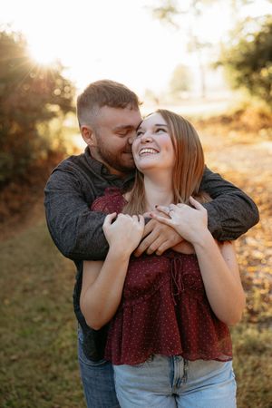 A couple embraces outdoors at golden hour, the man kissing her cheek as she smiles joyfully, wearing a red top and jeans.