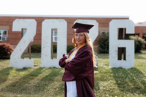 Young woman in maroon graduation cap and gown poses confidently in front of large 2026 yard letters outdoors.