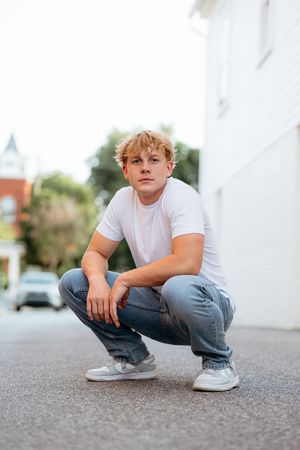 Young man in white t-shirt and jeans crouching on urban street, posing confidently for a casual outdoor photo.