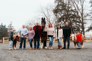 A group of people stands together with horses at a farm during winter, wearing casual clothing and holiday sweaters.