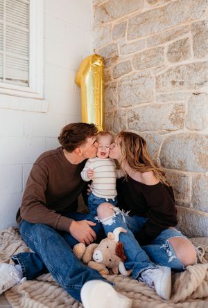 A loving couple sits together on a cushioned bench against a stone wall while their toddler plays with a stuffed toy.
